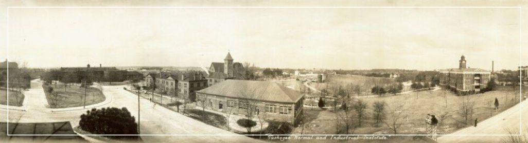 Wide panoramic view of a historic campus with buildings, trees, and a road or walkway under a pale sky