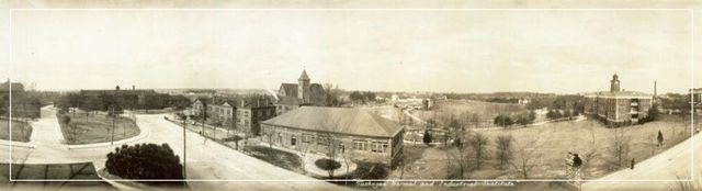 Wide sepia panorama of a town with buildings, roads, and open fields under a cloudy sky.