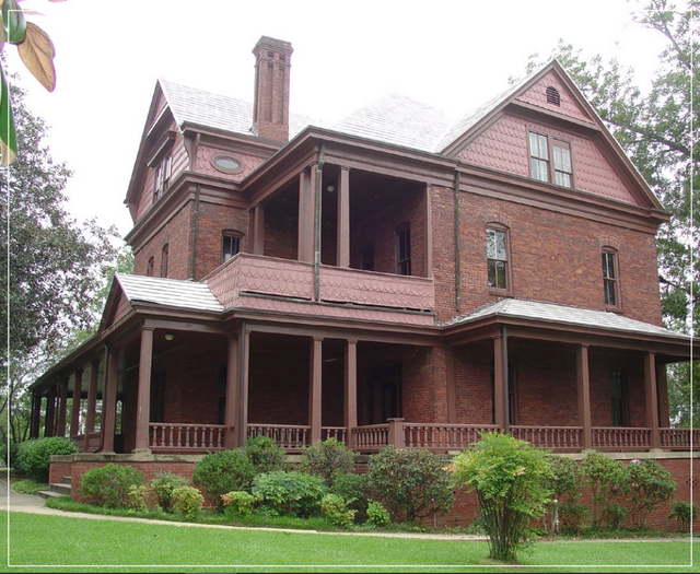 Victorian brick house with wraparound porch and gabled roof, surrounded by green shrubs and lawn.