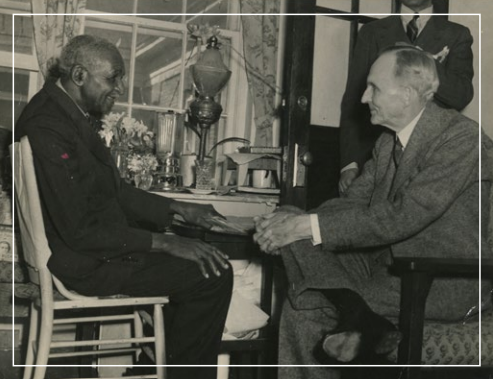 Two men seated facing each other in a room, one gesturing while talking beside a desk and lamp.