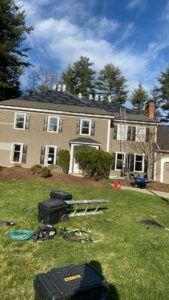 Two-story beige house with solar panels on roof. Tools and ladder in the yard, blue sky, and trees.