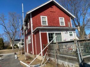 Red two-story house with siding. Scaffolding visible, part of house has protective wrap. Trees and fence in yard.
