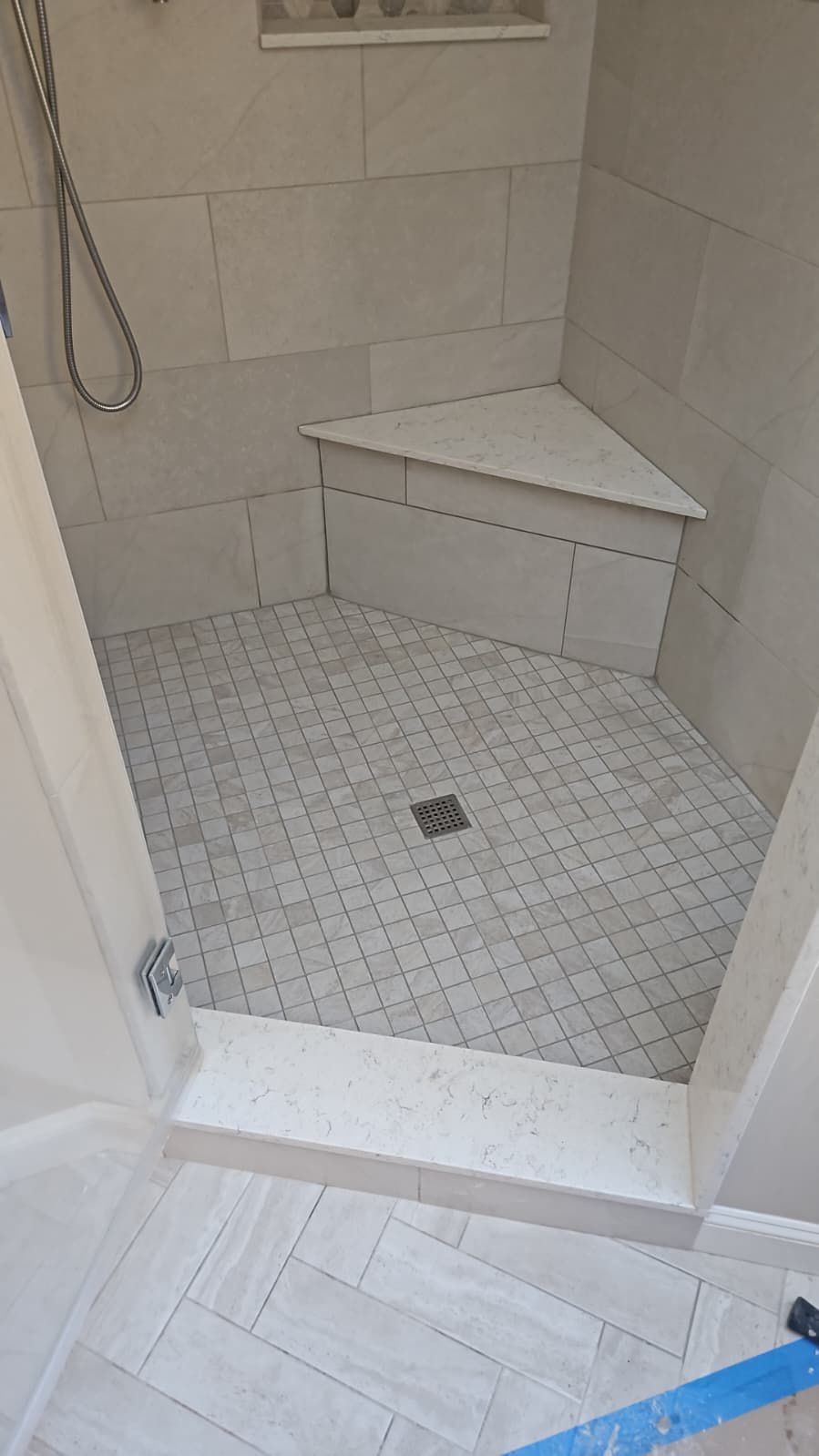 Shower stall with tiled walls, corner seat, and textured floor with a drain.