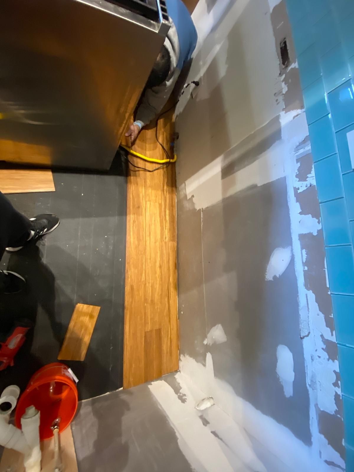 Installation of hardwood floor.  Person near partially installed flooring, adjacent to gray wall and blue tiled wall.