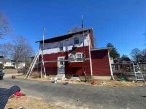 House exterior with scaffolding; worker repairs siding, blue sky.