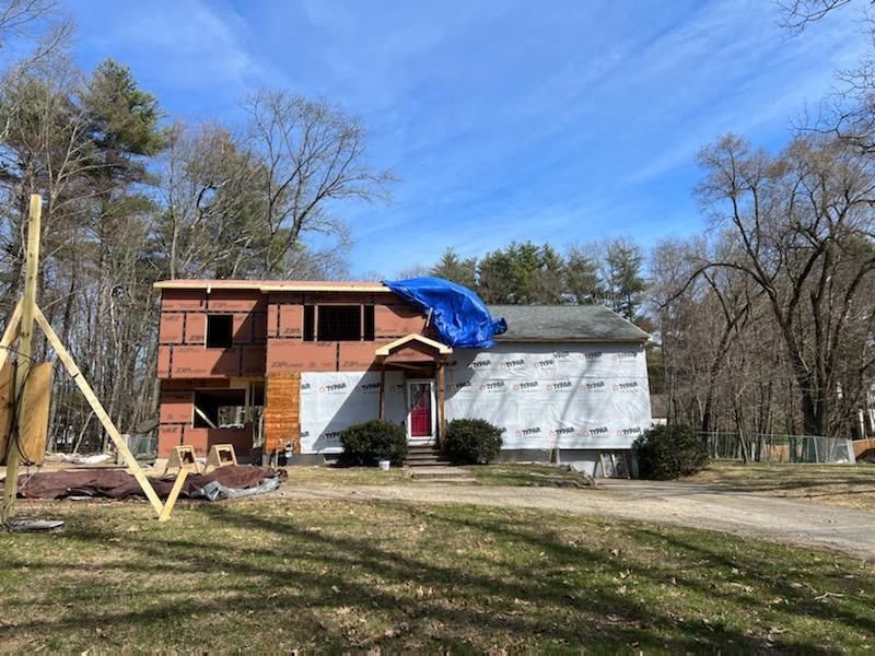 House under construction: unfinished walls, blue tarp on roof, surrounded by trees and grass, bright sky.