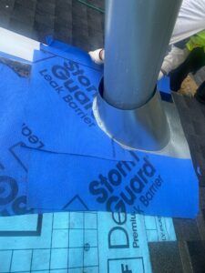 Blue roofing underlayment around a pipe on a dark shingle roof.