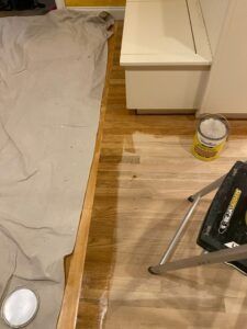 Wooden floor being refinished with paint, drop cloth protecting adjacent areas, paint can, and small step ladder.