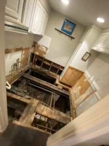 Kitchen with flooring removed, revealing subfloor and joists; white cabinets visible.