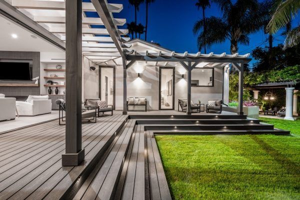 Nighttime shot of a modern home exterior with a wooden deck, pergola, and lit steps leading to a green lawn.