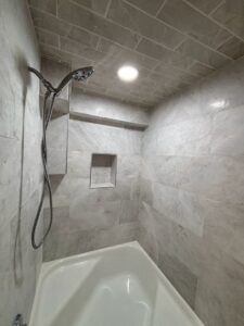 Bathroom with gray tiled walls, showerhead, recessed shelf, and a white tub.