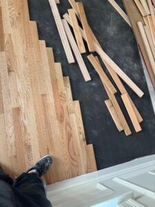 Wood flooring installation in progress; planks laid on concrete, worker's foot visible, white trim in the foreground.