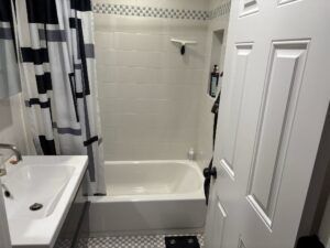 Small bathroom with white tiled shower/tub, sink, and closed white door. Black and white shower curtain.
