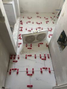White tile floor installation in progress, red leveling clips visible, with a fan in the center.