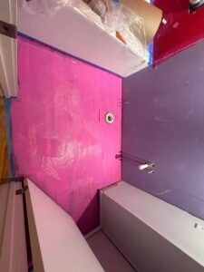 Pink waterproof membrane applied to a bathroom wall, with purple wall and white fixtures.