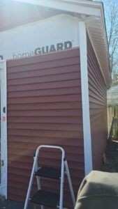 Siding installation on a building. Red horizontal siding, white trim, HomeGuard underlayment, and a stepladder are visible.