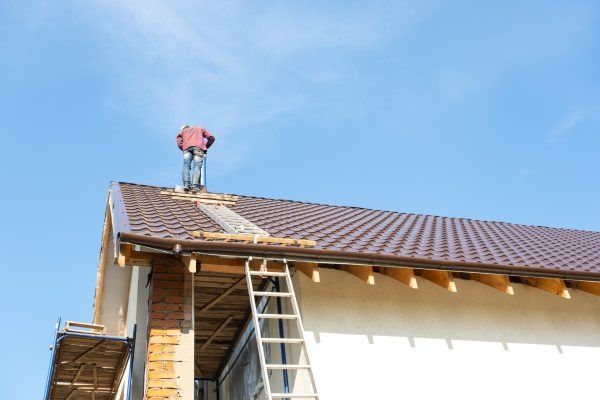 A person in red shirt on a roof working. A ladder leans against the building. Bright blue sky.
