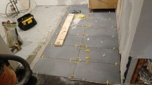 Floor tile installation in progress. Gray tiles with yellow spacers on a concrete floor. Tools and materials nearby.