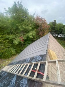Metal roof installation with ladder, partially completed, forest background.