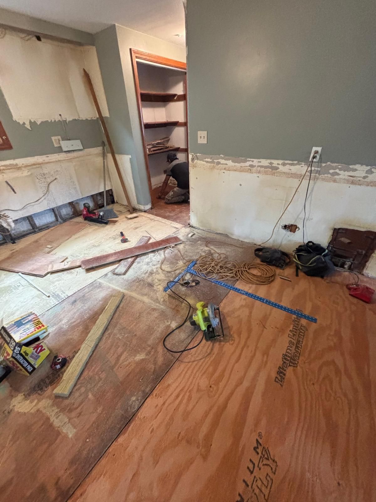 Room undergoing renovation; plywood on floor, exposed wall, tools, person working in a doorway.