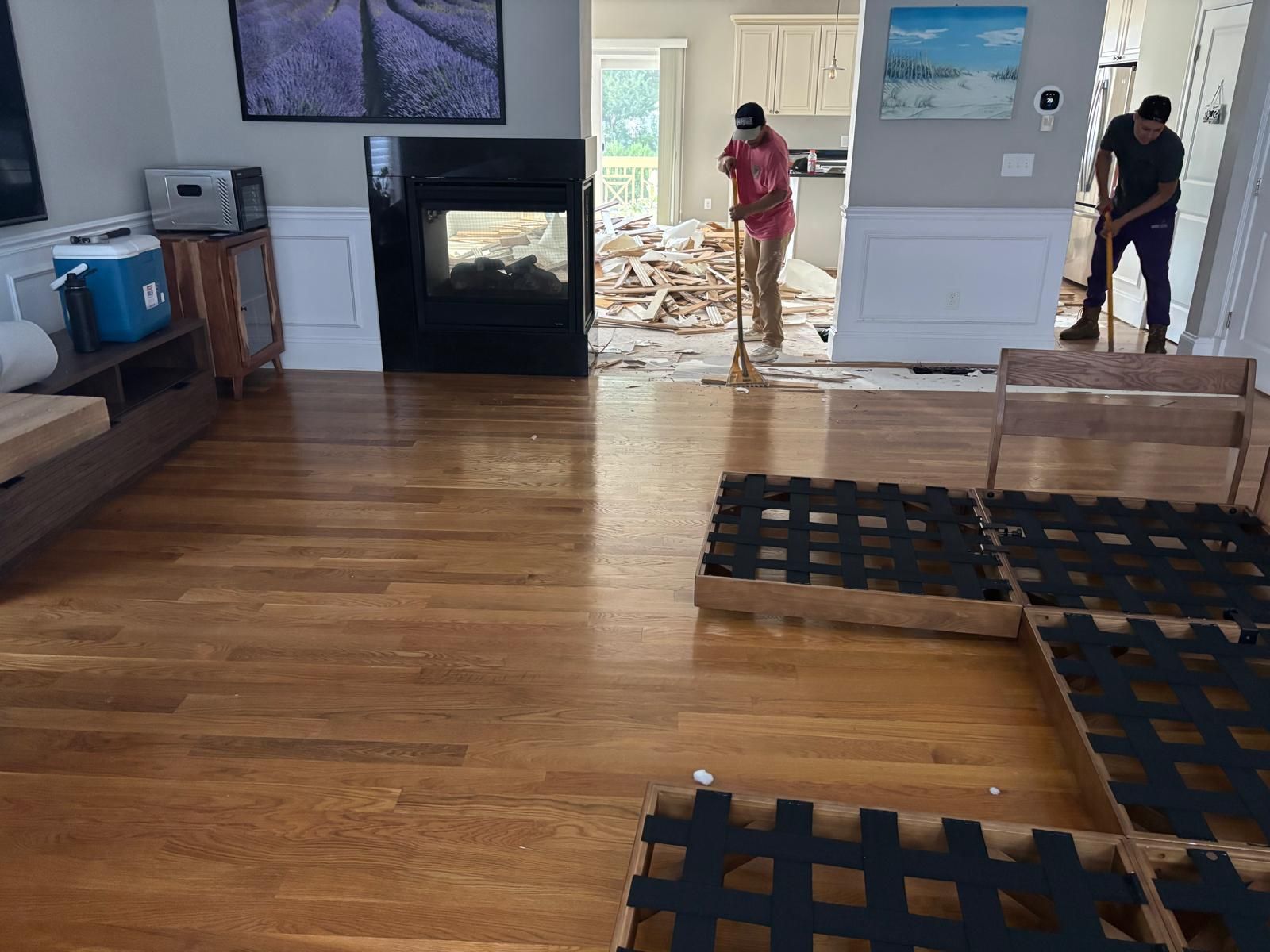 People cleaning debris from a room with hardwood floors. One sweeps, another gathers. Fireplace and open kitchen visible.