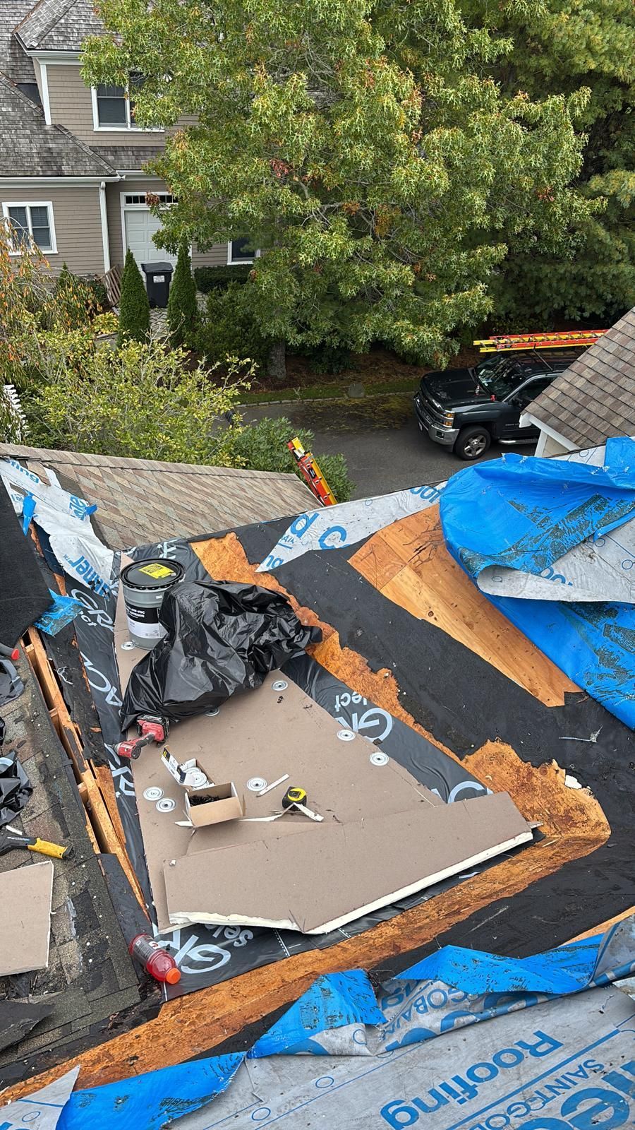 Rooftop with blue tarp and debris. Partially removed shingles, tools, and a black garbage bag are visible.