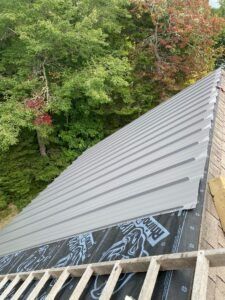 Metal roofing panels being installed on a roof with a forest background. Gray panels, wood, and underlayment visible.