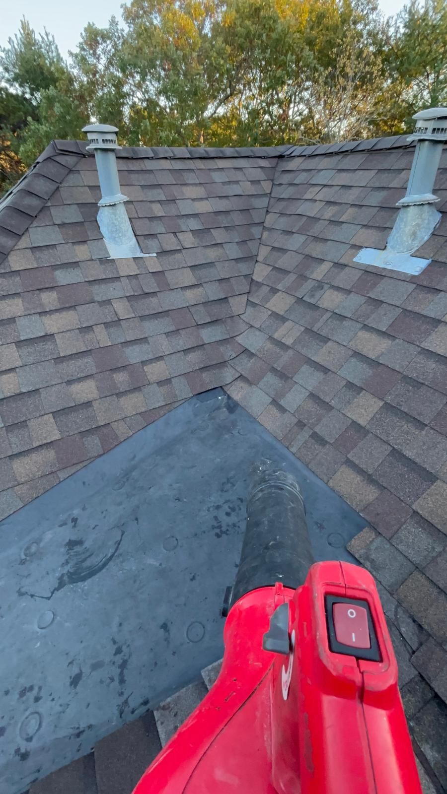 Red leaf blower on a dark roof corner, blowing debris. Brown shingles and vents in background.