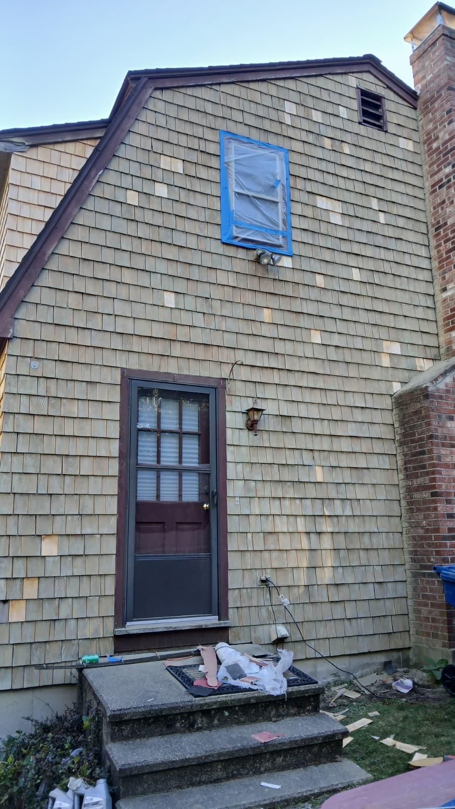 Exterior of a house with cedar shingle siding, a door, a window covered in blue tape, and a brick chimney.