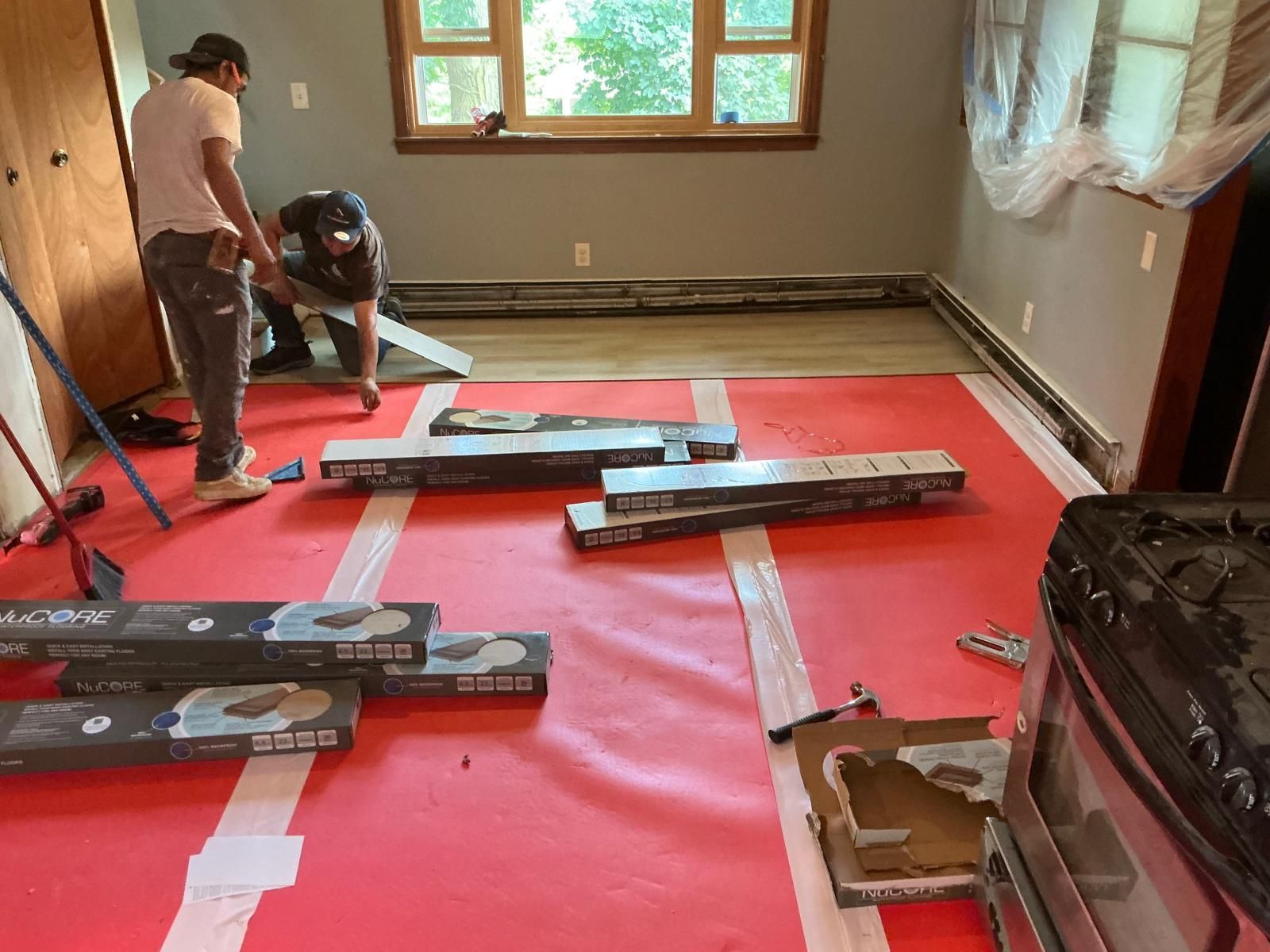 Two workers installing flooring in a room. Boxes of flooring on red underlayment. Window in the background.