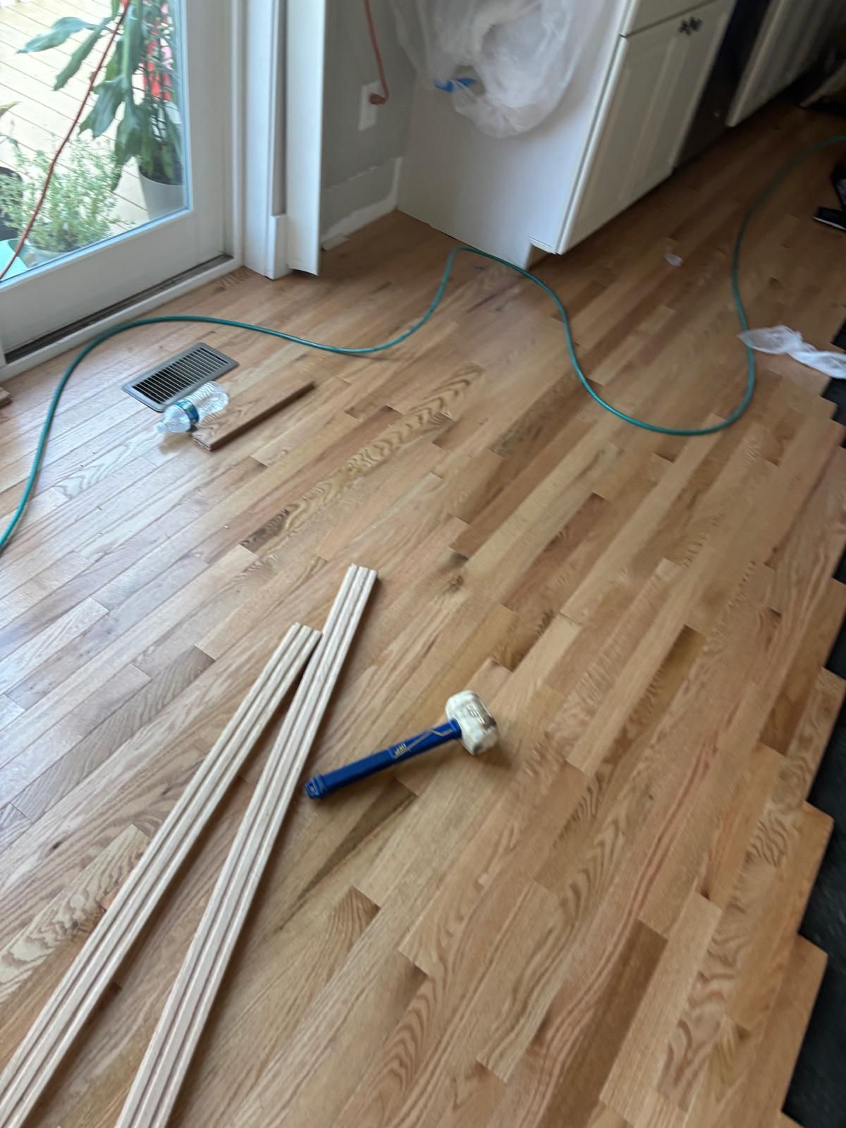 Wood flooring installation in progress; includes boards, hammer, trim, and hose.