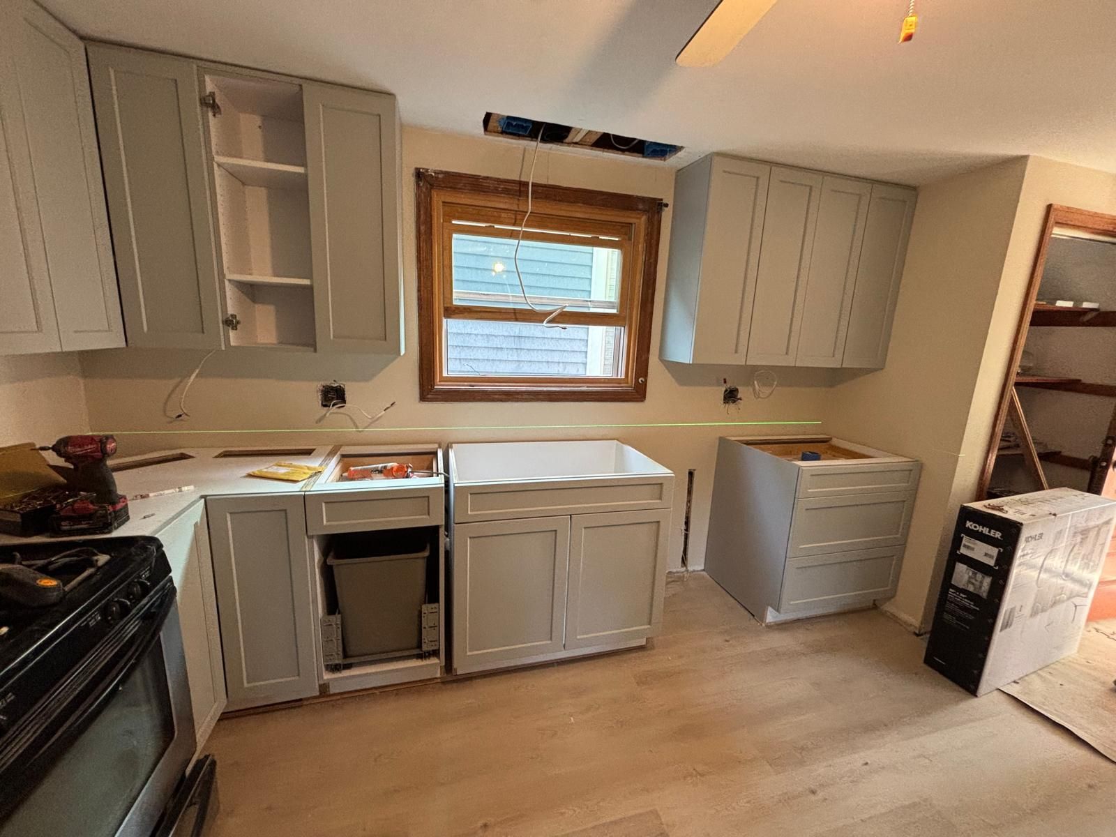 Kitchen under renovation: light gray cabinets, white sink, bare wood floors, window, oven.