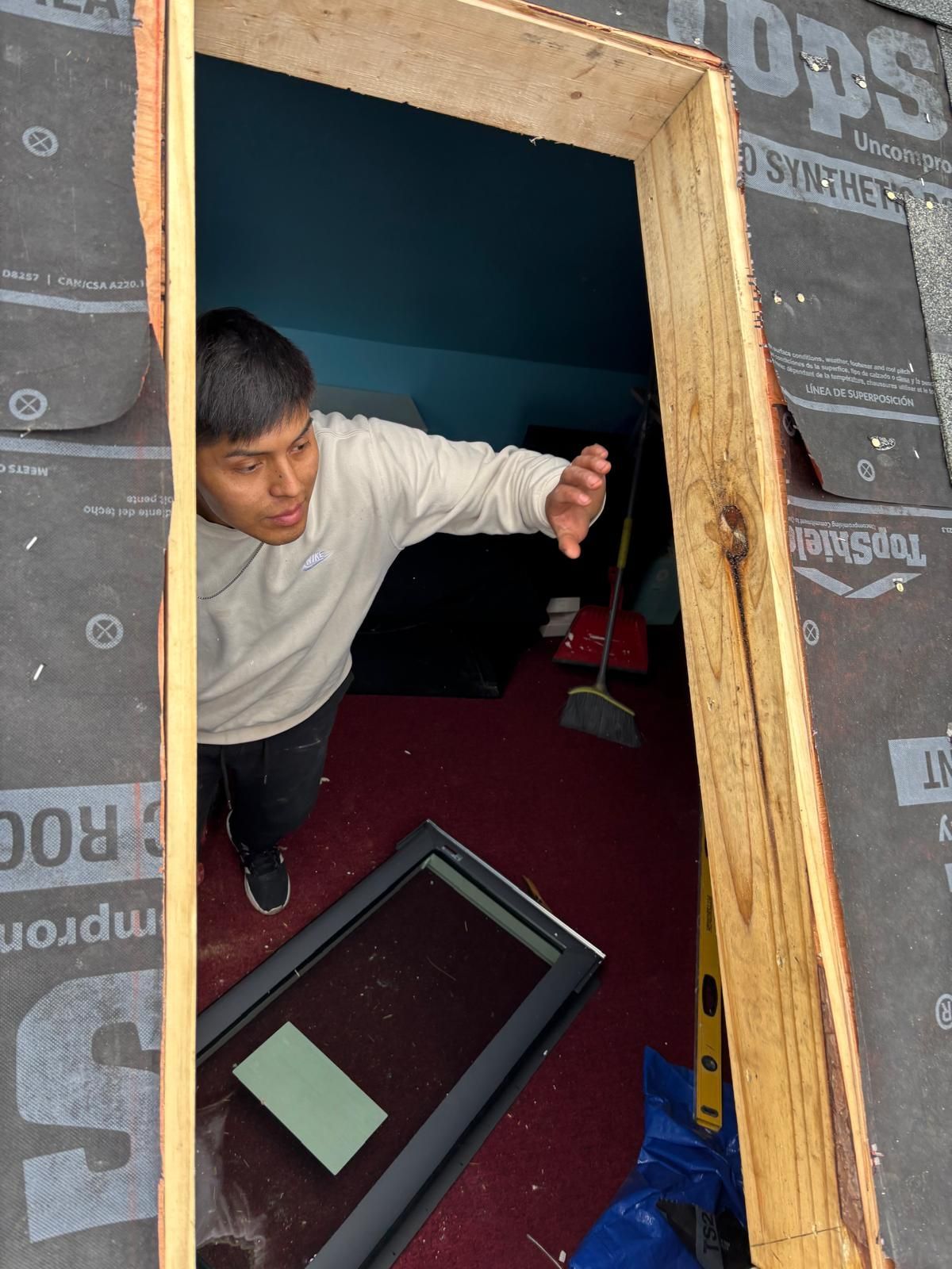 Man reaching from rooftop opening; window and tool inside. Interior, red rug, blue wall.