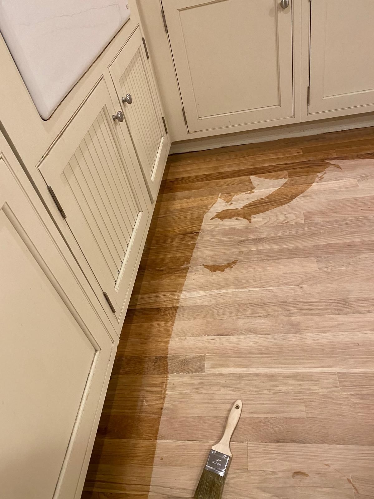 Applying stain to wooden floor with a brush; cabinets on left.