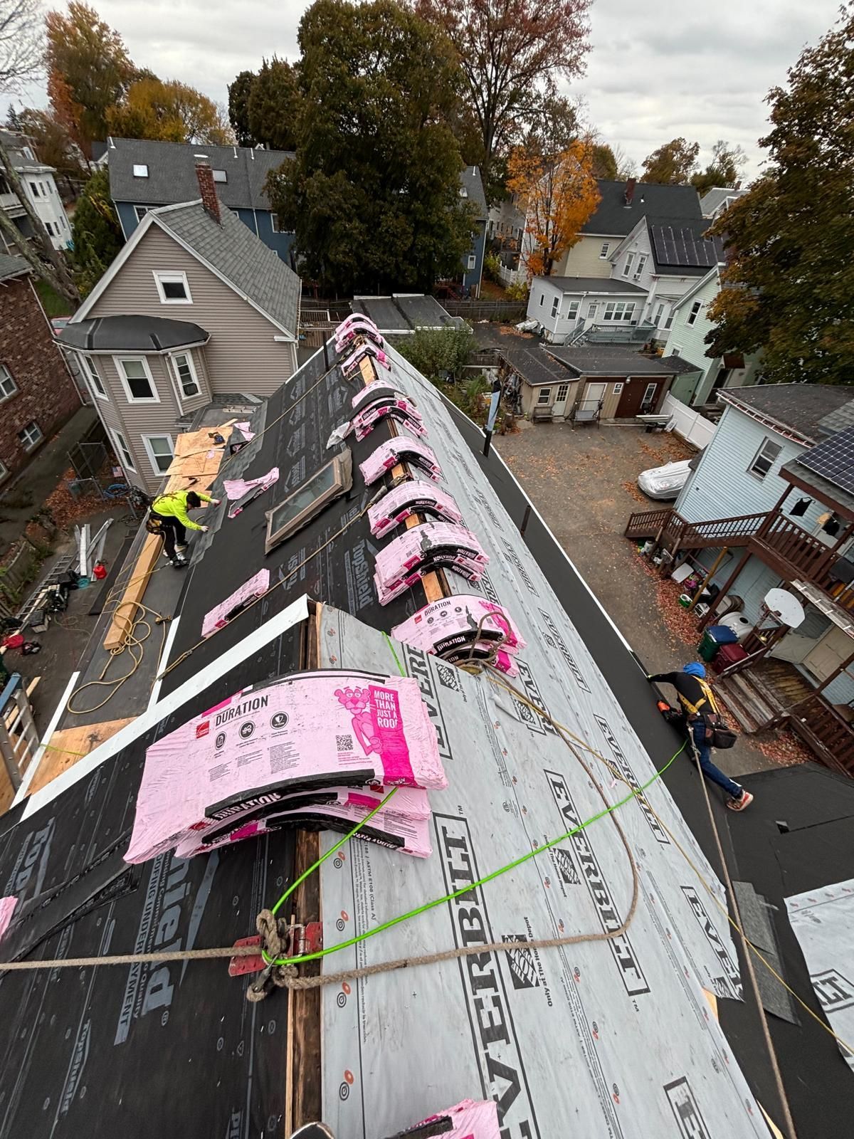 Roof construction: workers on a steep roof with black underlayment, pink insulation, and safety harnesses.