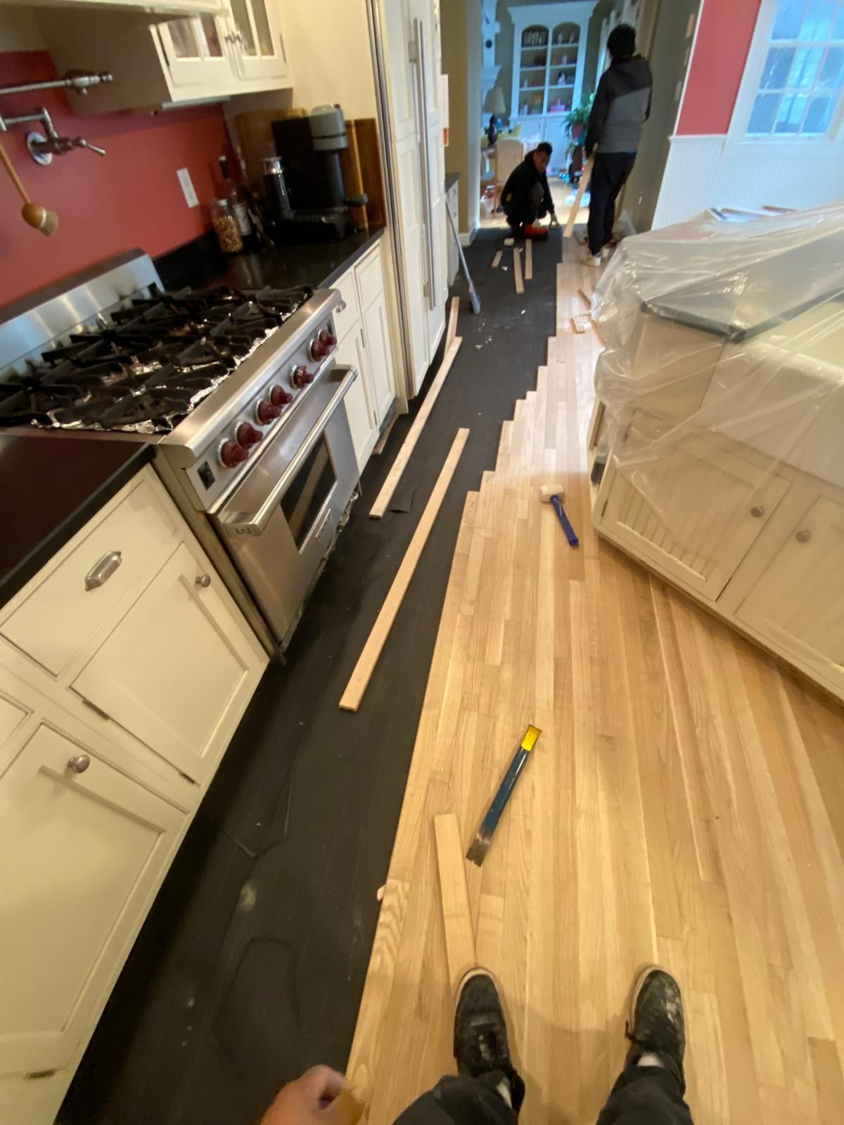 Kitchen with partially installed hardwood flooring; workers install planks near cabinets and stove.