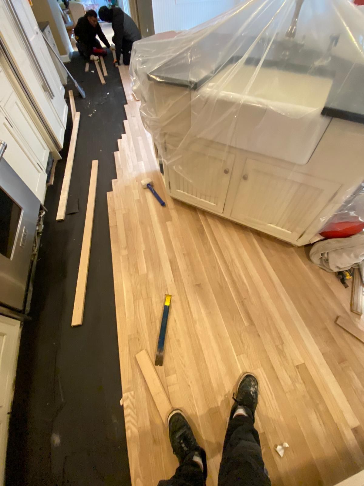 Wood floor installation in progress: installers, tools, black sheeting, and cabinets covered in plastic.