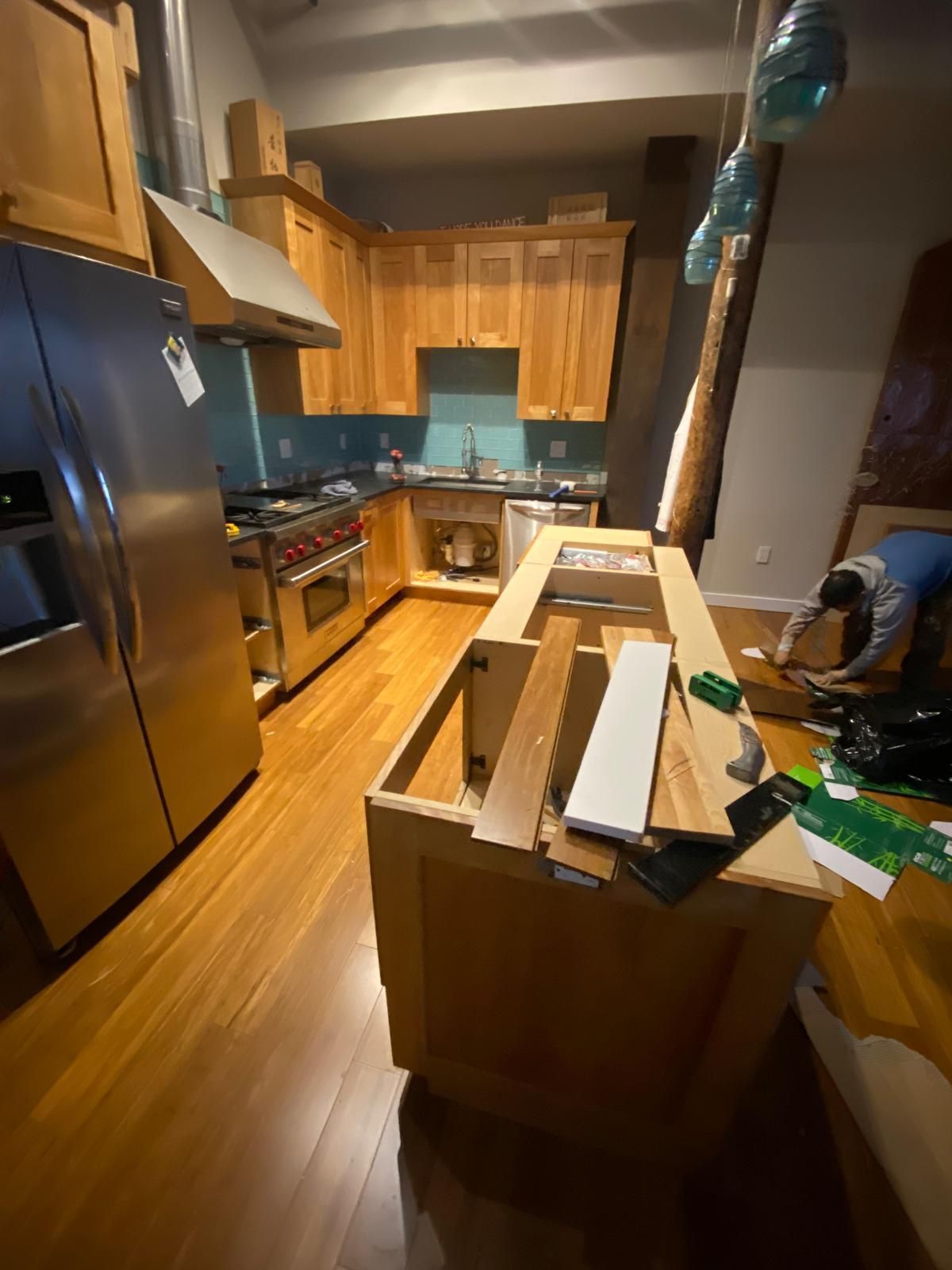 Kitchen with wooden cabinets and island under construction. Stainless steel appliances, blue backsplash, and bamboo flooring.