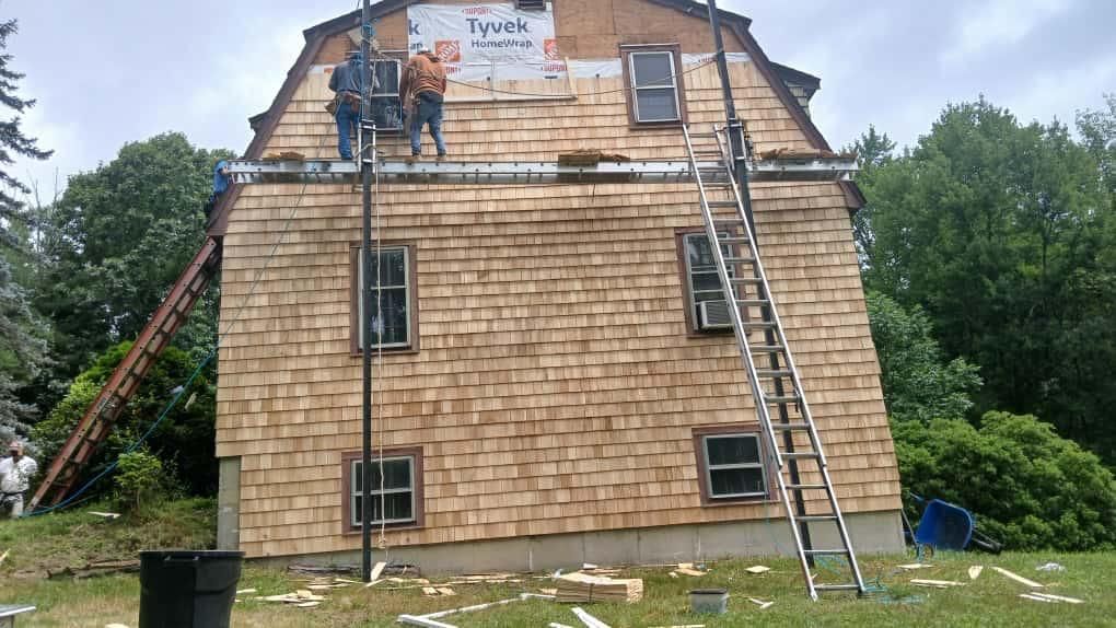 Two roofers on a house with wood shingles, installing roofing. Ladders and tools visible.