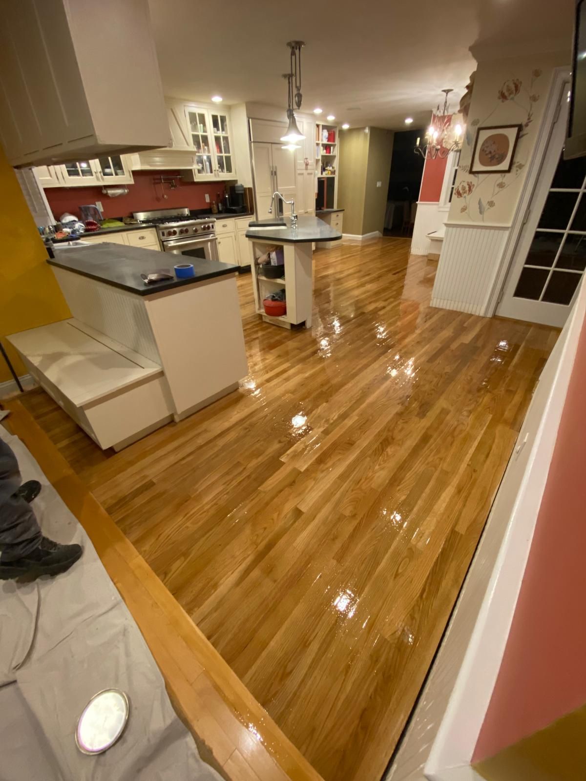 Shiny hardwood floors in a kitchen, island with a dark countertop, white cabinets and red backsplash.
