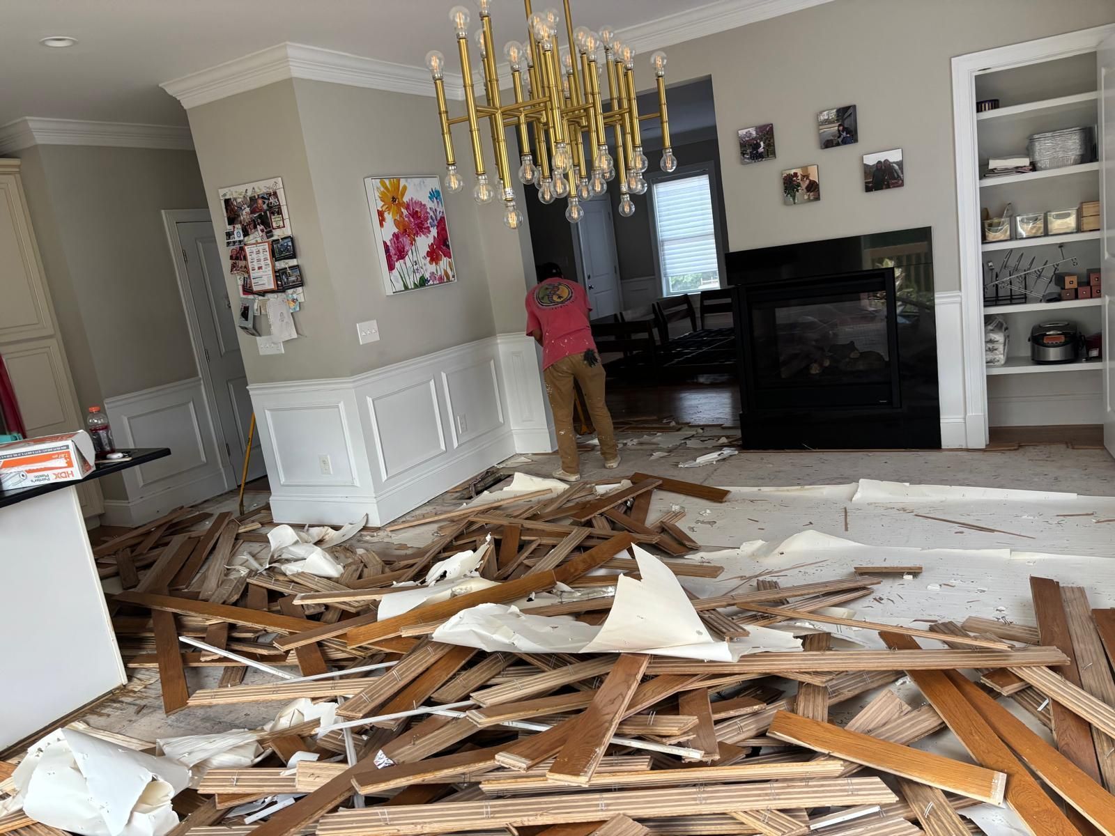 Room undergoing flooring renovation, debris scattered. Person standing, a chandelier hangs, cabinets and fireplace visible.
