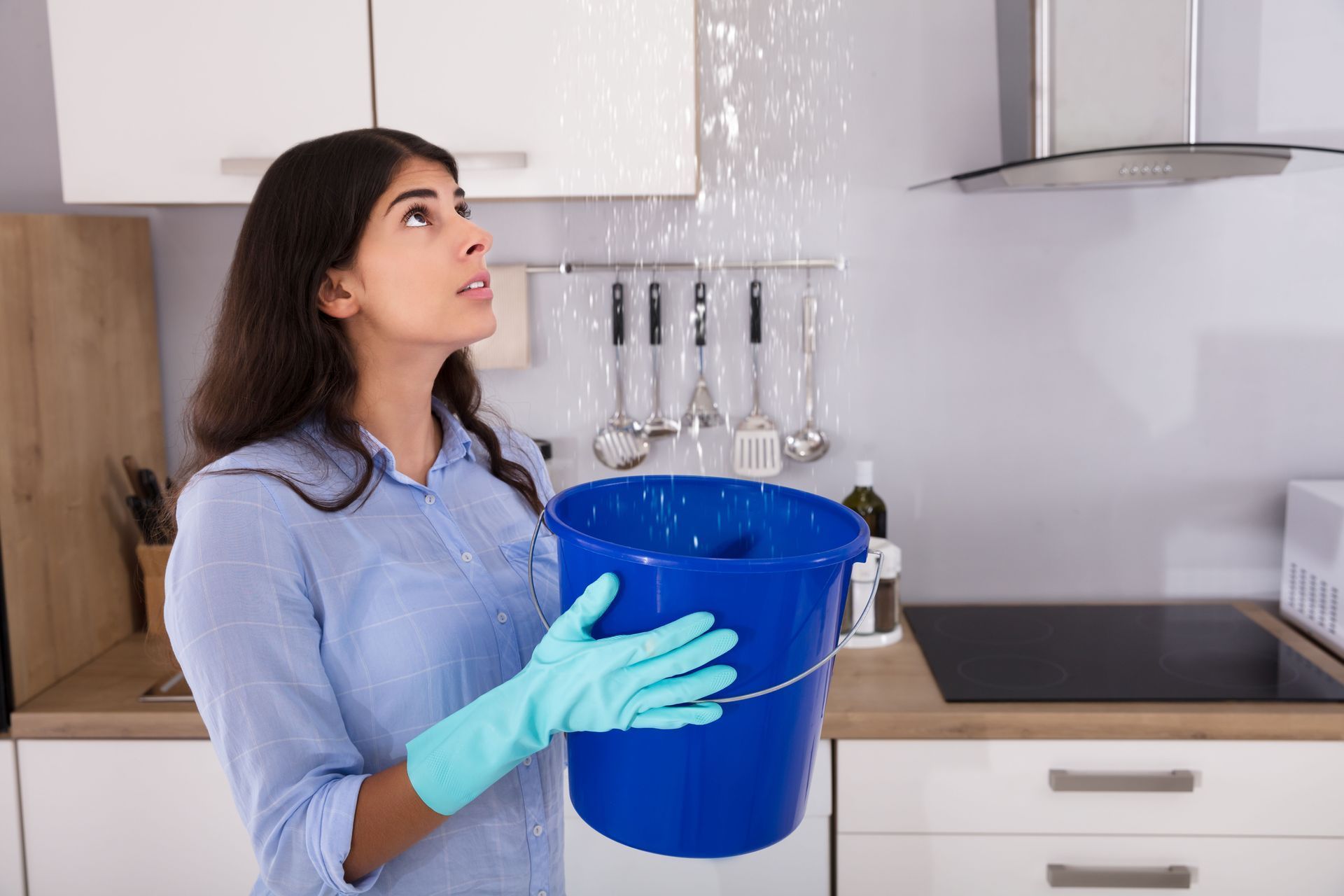 Woman in kitchen holding bucket, looking up at ceiling leak. Blue bucket, water falling.