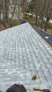Gray shingle roof, partially completed, with a building and trees in the background.