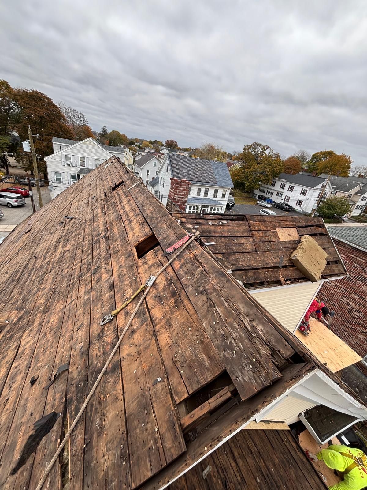 Damaged roof with missing sections; worker nearby, overcast sky, residential area in background.