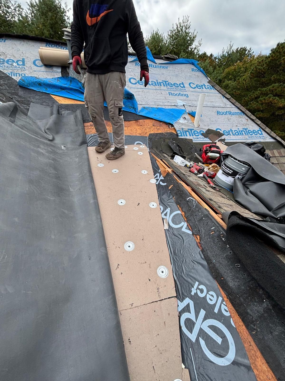 Roofer standing on a partly re-roofed section, wearing casual clothes and gloves, cloudy day.