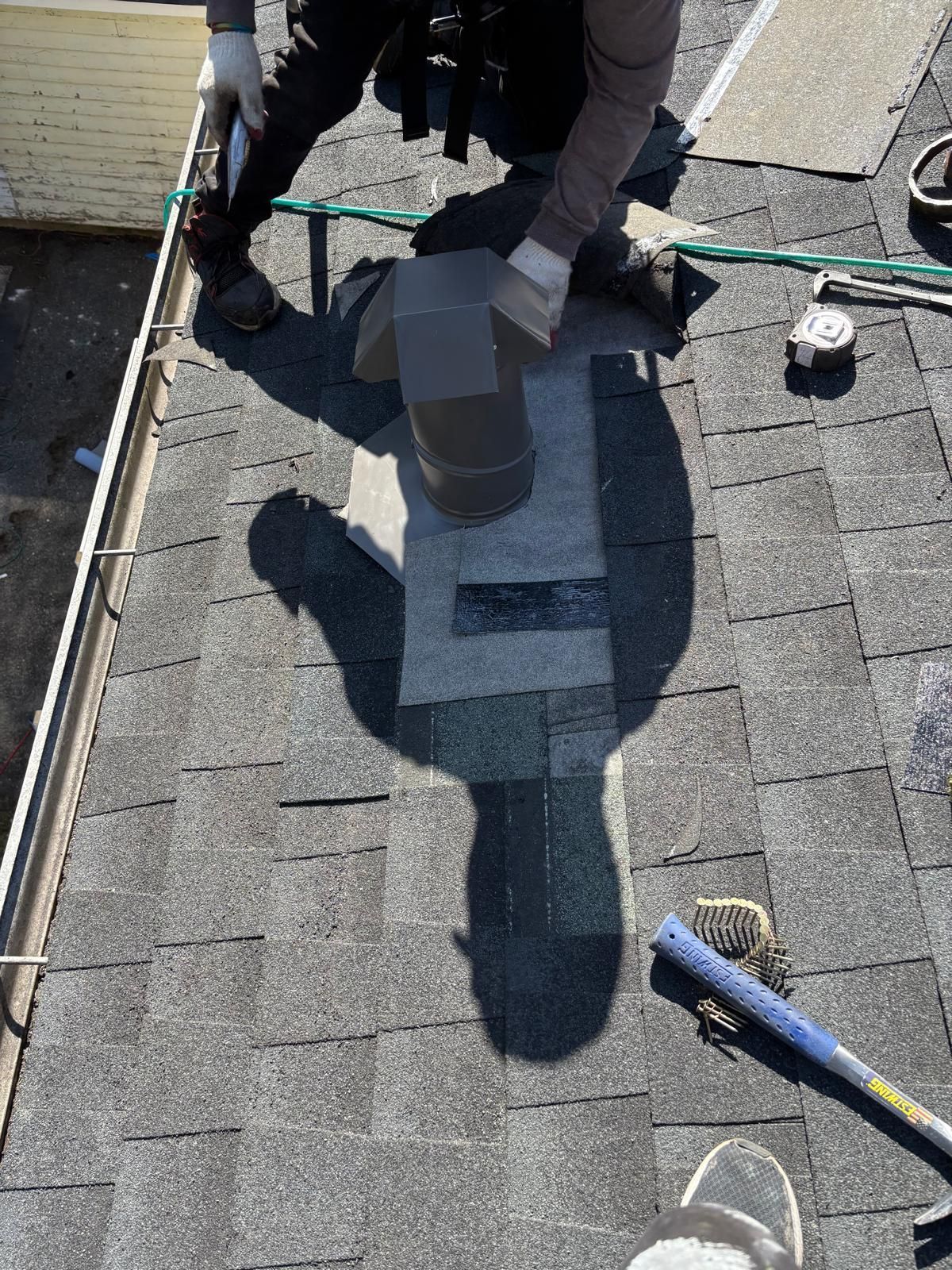 Roofer installing a chimney flashing on a shingled roof, outdoors. Person kneels, holding materials and tools.