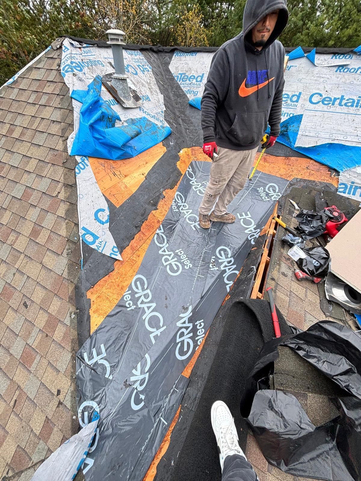 A person standing on a roof is applying a black membrane. Brown shingles and blue tarps are visible.