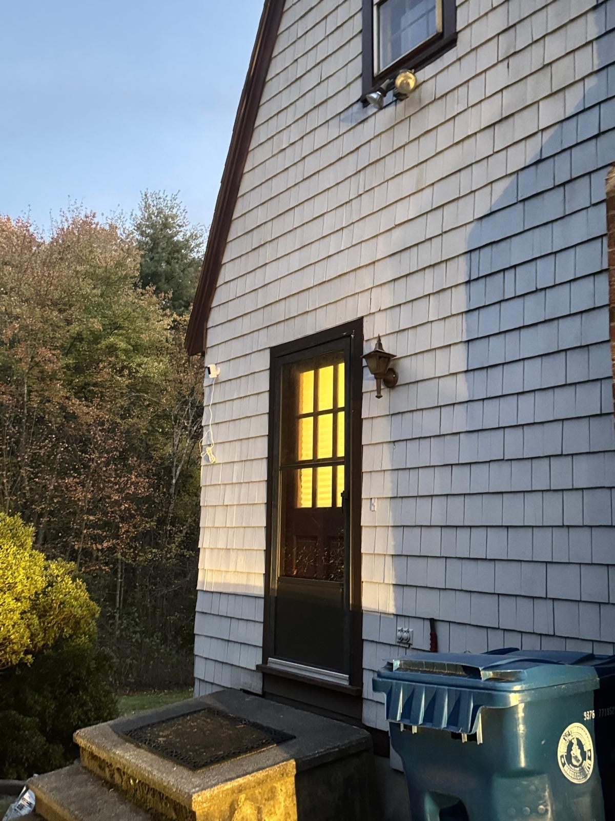 Exterior of a house with gray shingle siding, a black door with sunlight shining through, and a blue trash can.