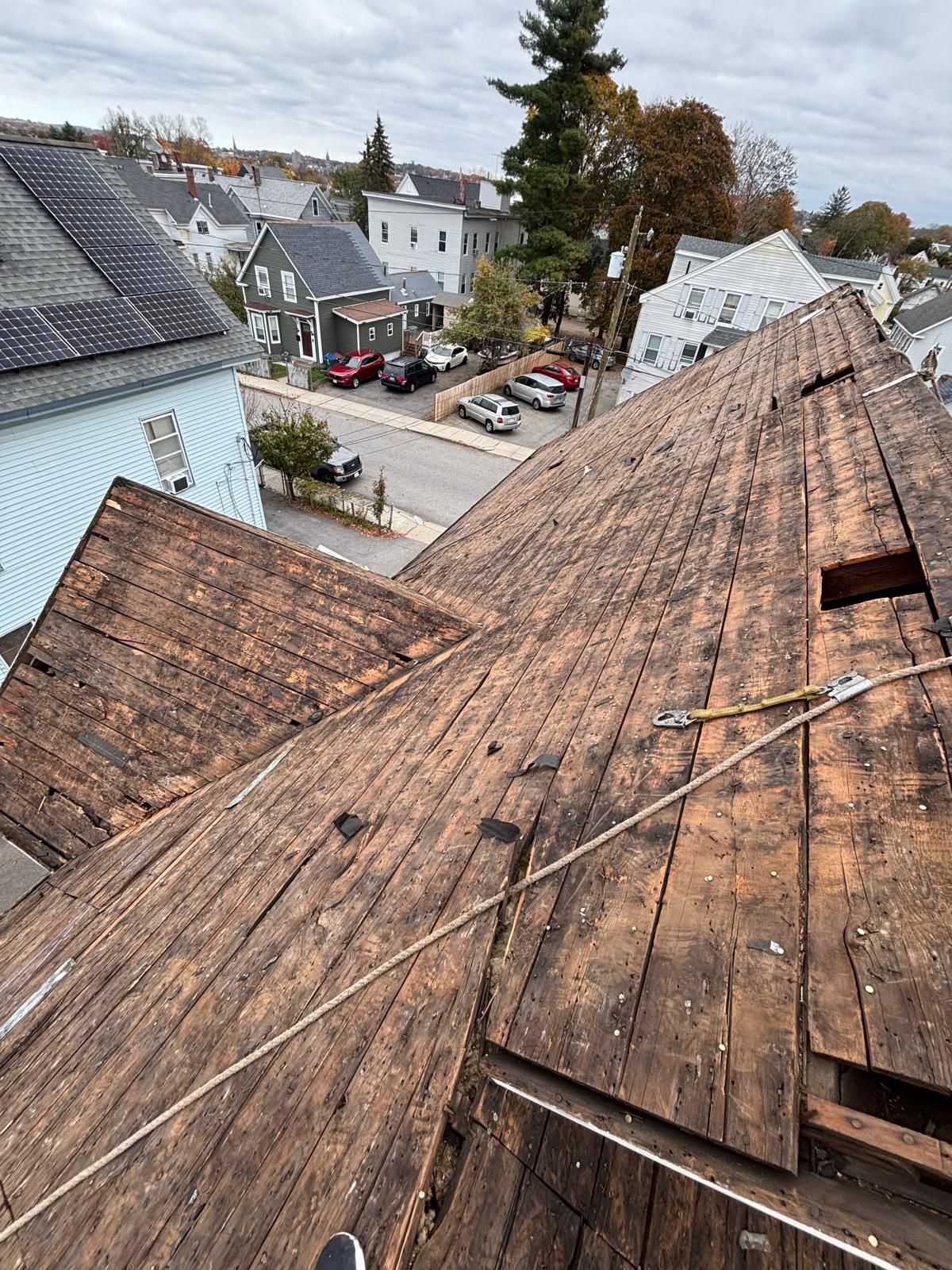 View of a weathered, deteriorating roof with damaged wooden shingles. Neighborhood in the background on a cloudy day.