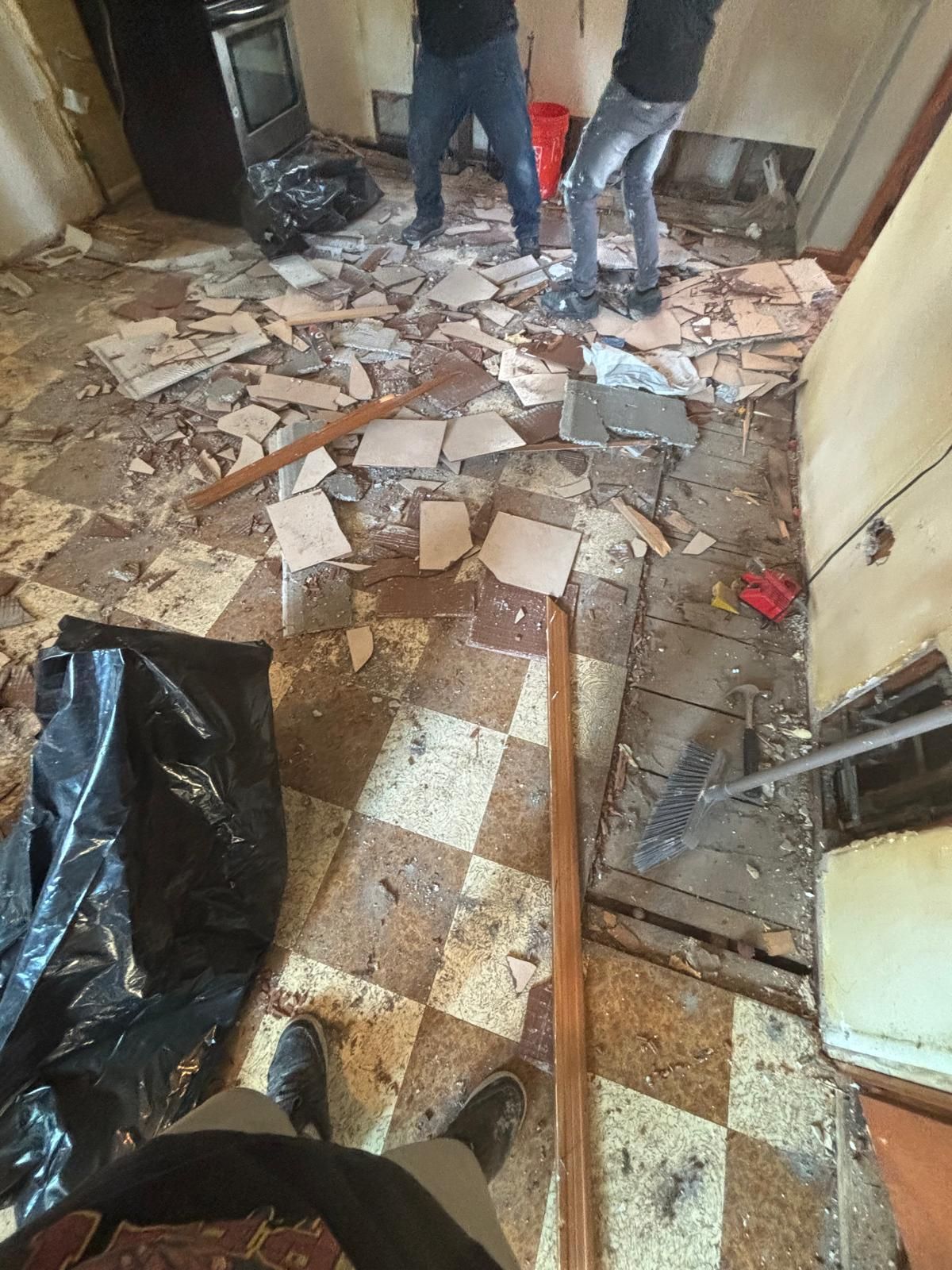 Two people removing flooring in a room. Debris covers the floor, and a stove is visible.